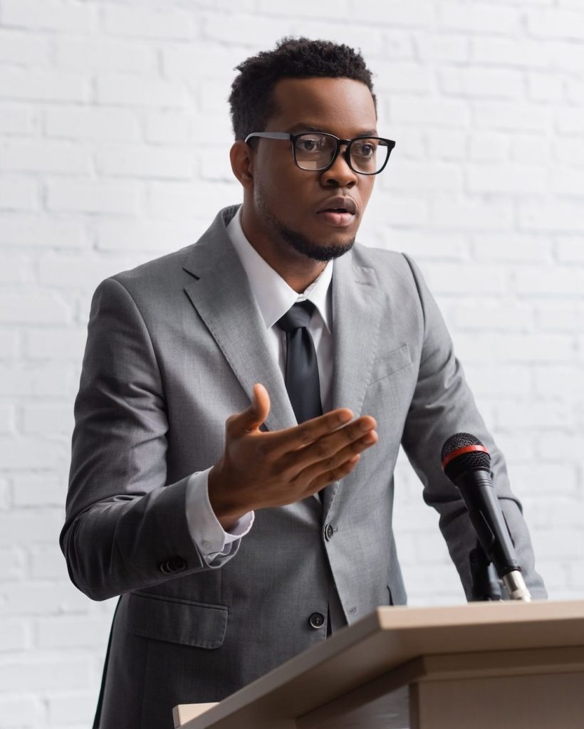 confident-african-american-business-speaker-on-tribune-with-microphone-in-conference-hall-e1661098107411-ptlj7h09cn84dvz3nhviu4i6zy13ue3aqbqy2sxhxc